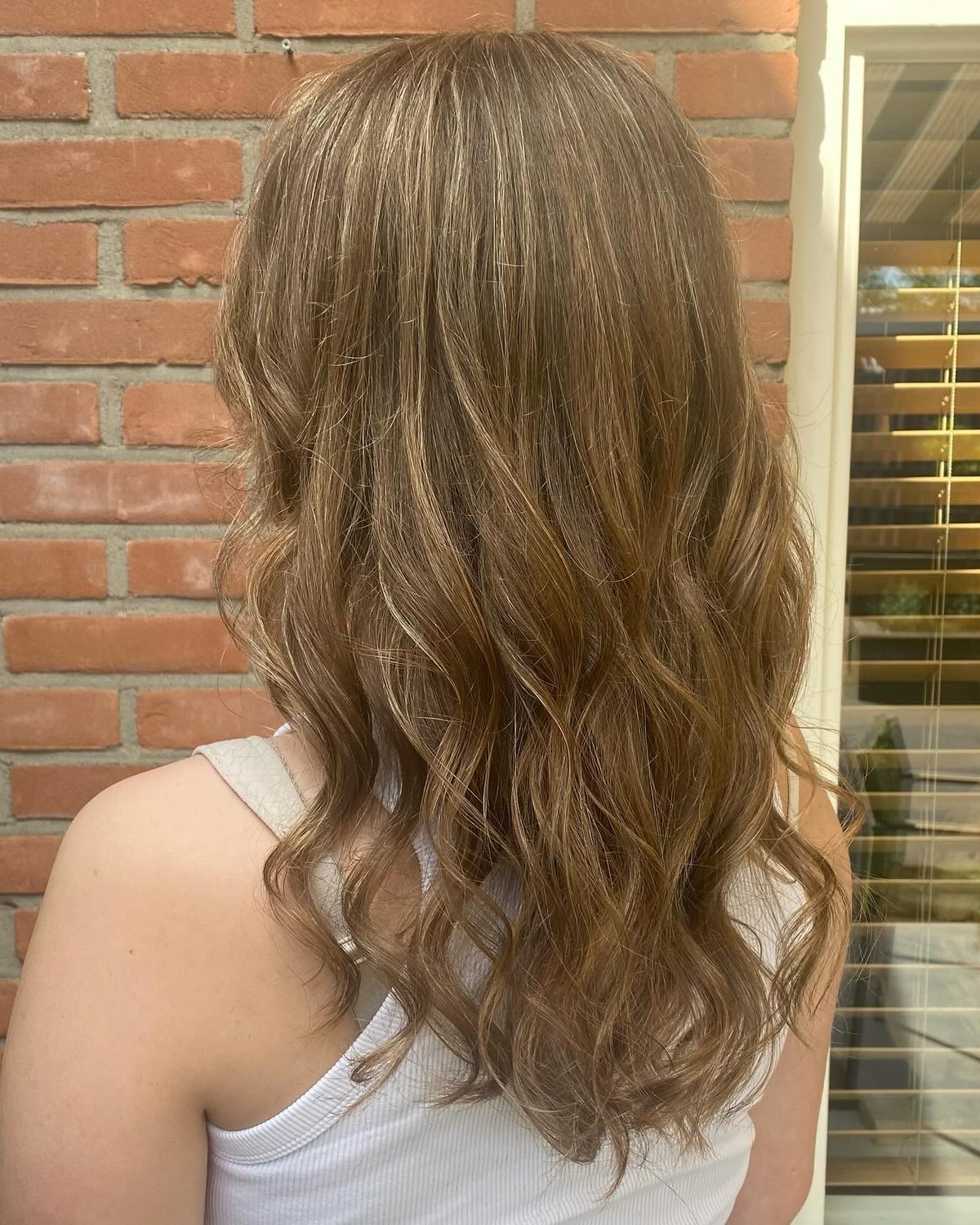 Lichtbruin haar met highlights en losse golven, buiten tegen een bakstenen muur